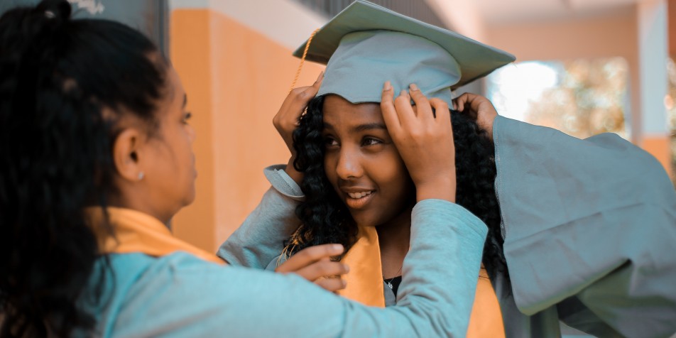An image of two women in graduation gowns. One helps the other adjust their cap.