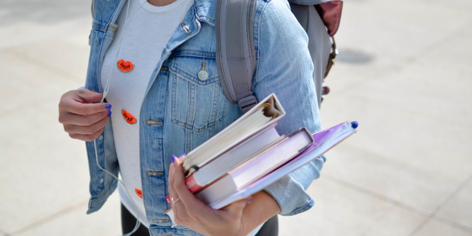 Image of young woman in jean jacket, backpack and headphones, carrying textbooks. 